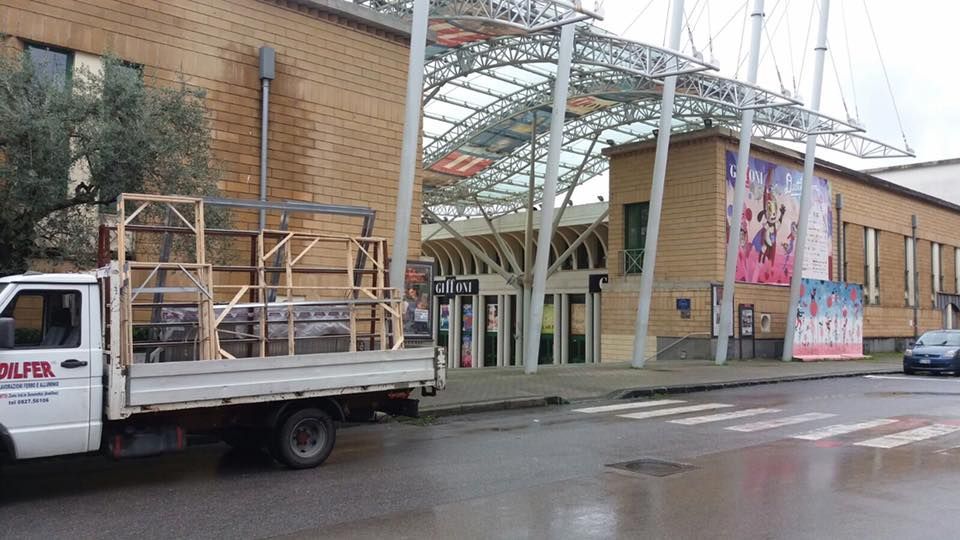 Un camion bianco parcheggiato davanti a un edificio beige con un ingresso in vetro e grandi poster alle pareti. È una giornata nuvolosa.