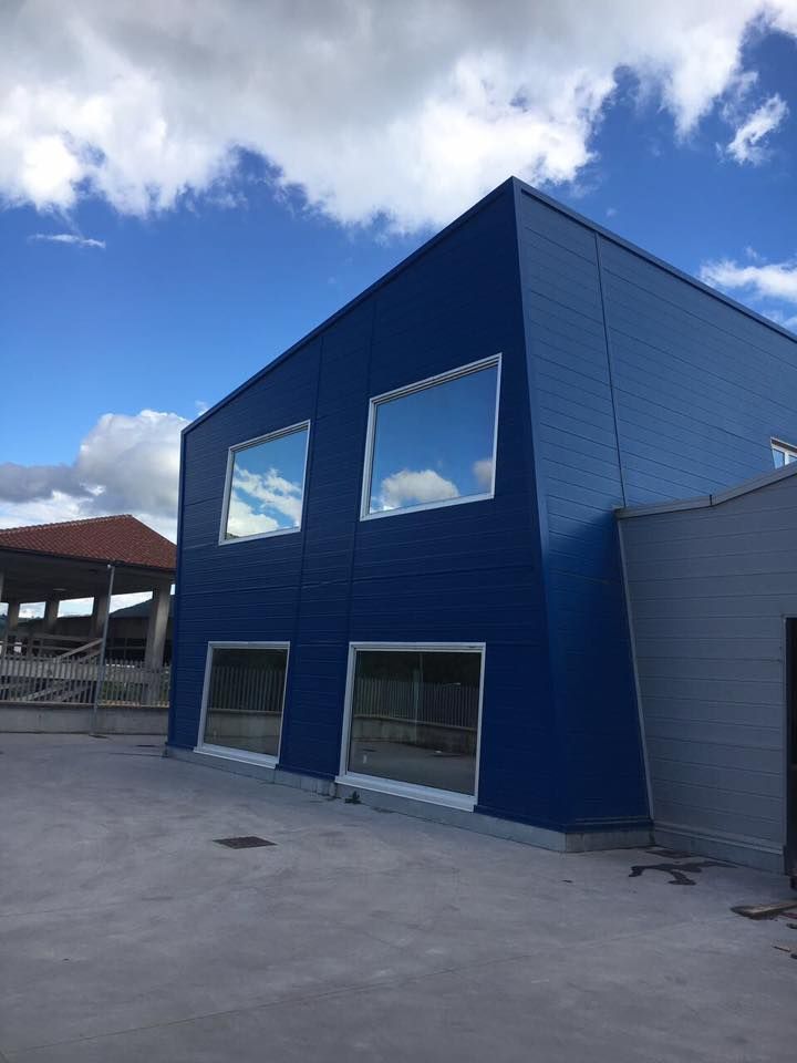 Edificio blu con finestre rettangolari che riflettono il cielo. Pavimento in cemento e una struttura grigia parzialmente visibile sullo sfondo.