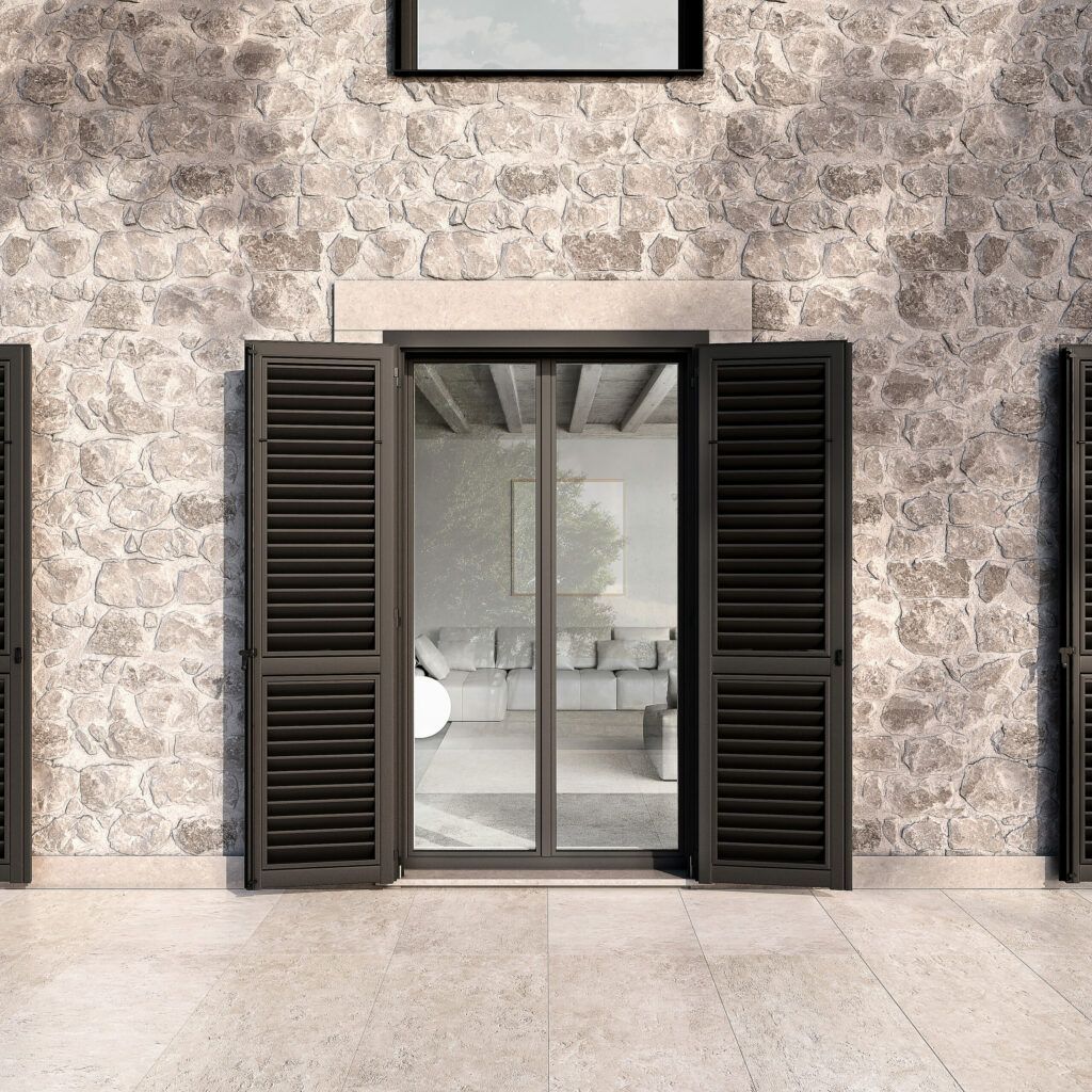 Vista esterna di un edificio con muri in pietra e persiane scure aperte che rivelano un soggiorno.