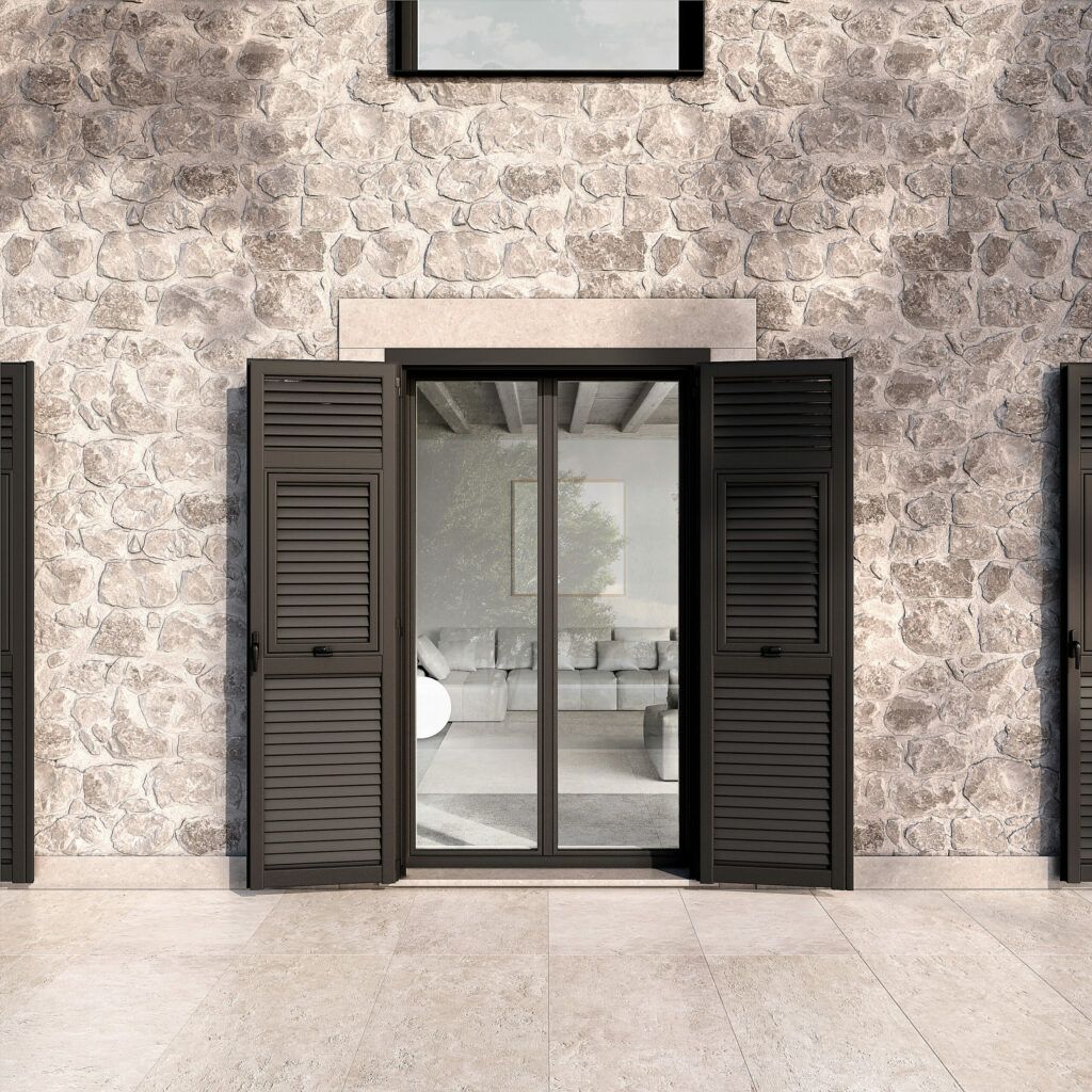 Ripresa esterna di un edificio con persiane nere che incorniciano una porta a vetri, rivelando un soggiorno; sullo sfondo un muro di pietra grigia.