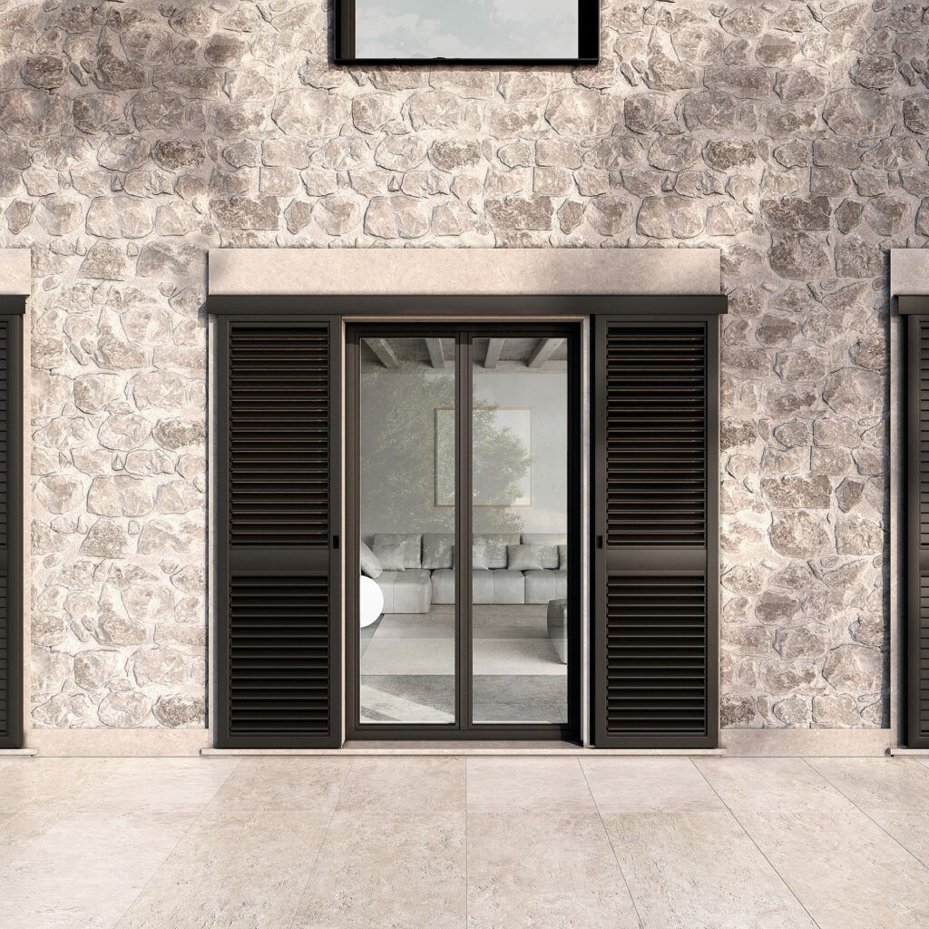 Vista esterna di un edificio in pietra con una porta scorrevole in vetro fiancheggiata da persiane scure. La porta conduce a un soggiorno ben illuminato.