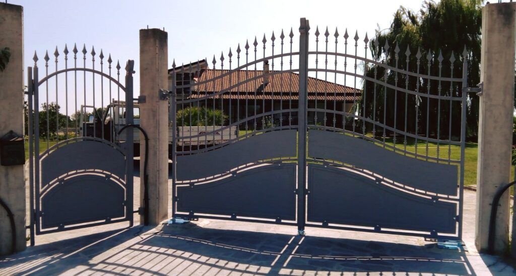 Cancello d'accesso in metallo grigio con motivo decorativo curvo, fiancheggiato da pilastri in cemento. Sullo sfondo, una casa con il tetto marrone.
