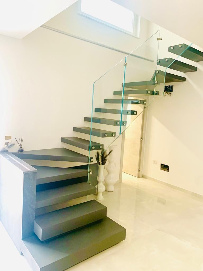 Scala moderna con gradini grigi e ringhiera in vetro contro una parete bianca e un pavimento lucido.