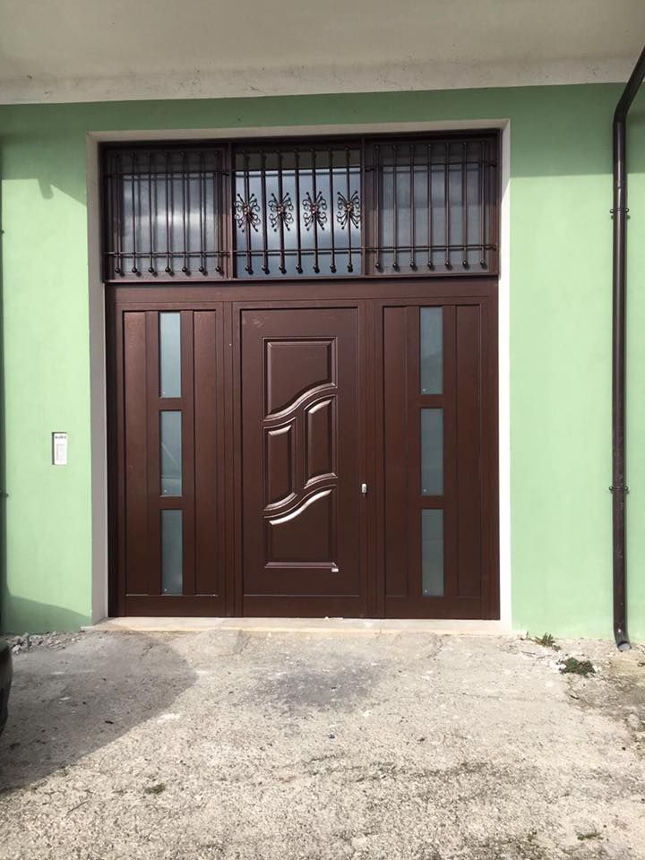 Porta in metallo marrone con pannelli in vetro smerigliato e decorazioni in ferro battuto nella parte superiore, appoggiata a una parete verde.