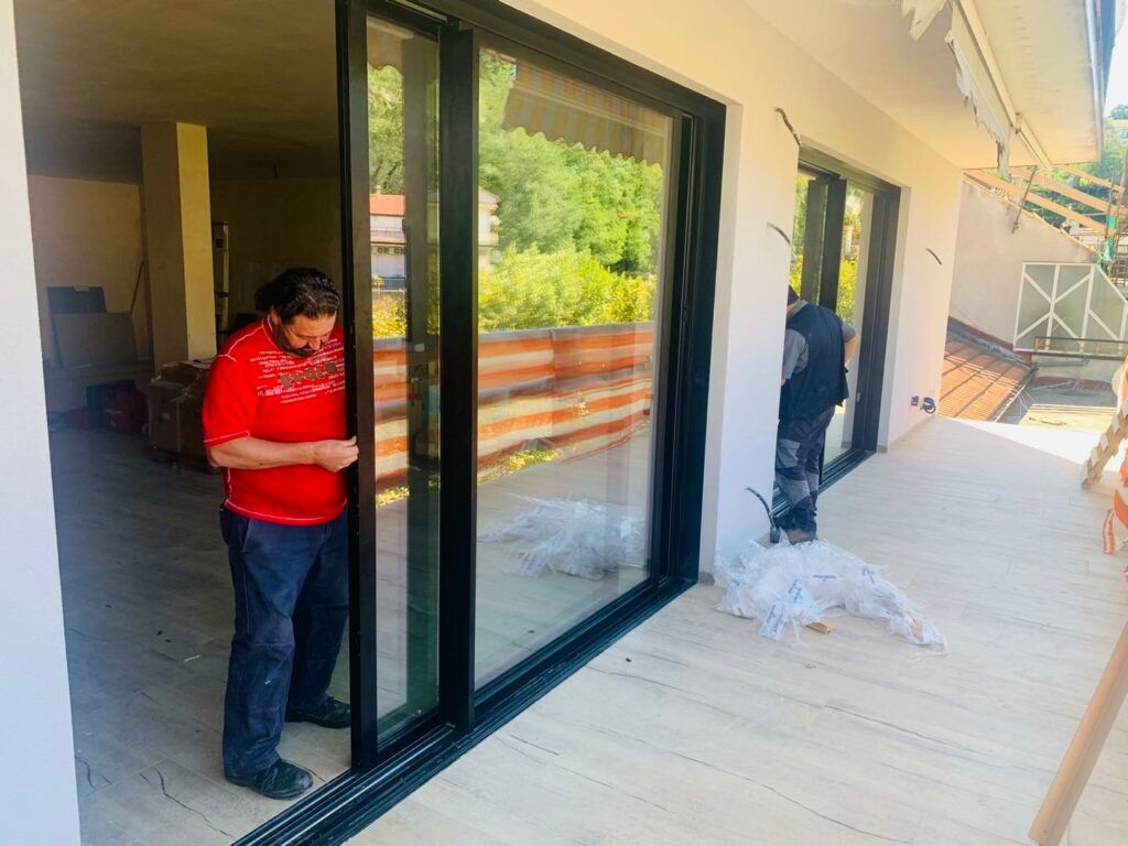 Un uomo con una camicia rossa sta aggiustando una porta scorrevole in vetro. Un'altra persona lavora lì vicino, su un balcone con vista sul verde.