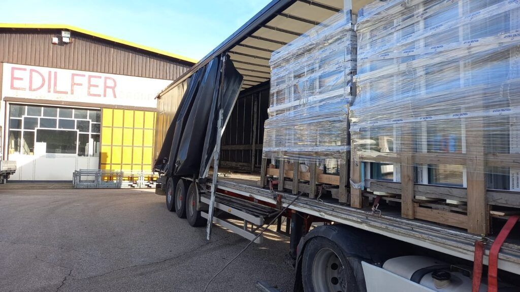 Un autoarticolato è carico di vetri presso un edificio EDILFER. Il telone del camion è aperto, rivelando il carico.