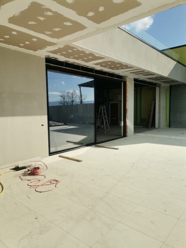 Un cantiere con una grande porta scorrevole in vetro con cornice nera. L'interno è grezzo, con cartongesso e struttura a vista.