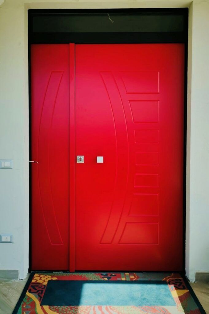 Porta d'ingresso rossa con un piccolo pannello laterale, incorniciata da una cornice nera contro una parete bianca. Davanti c'è uno zerbino colorato.