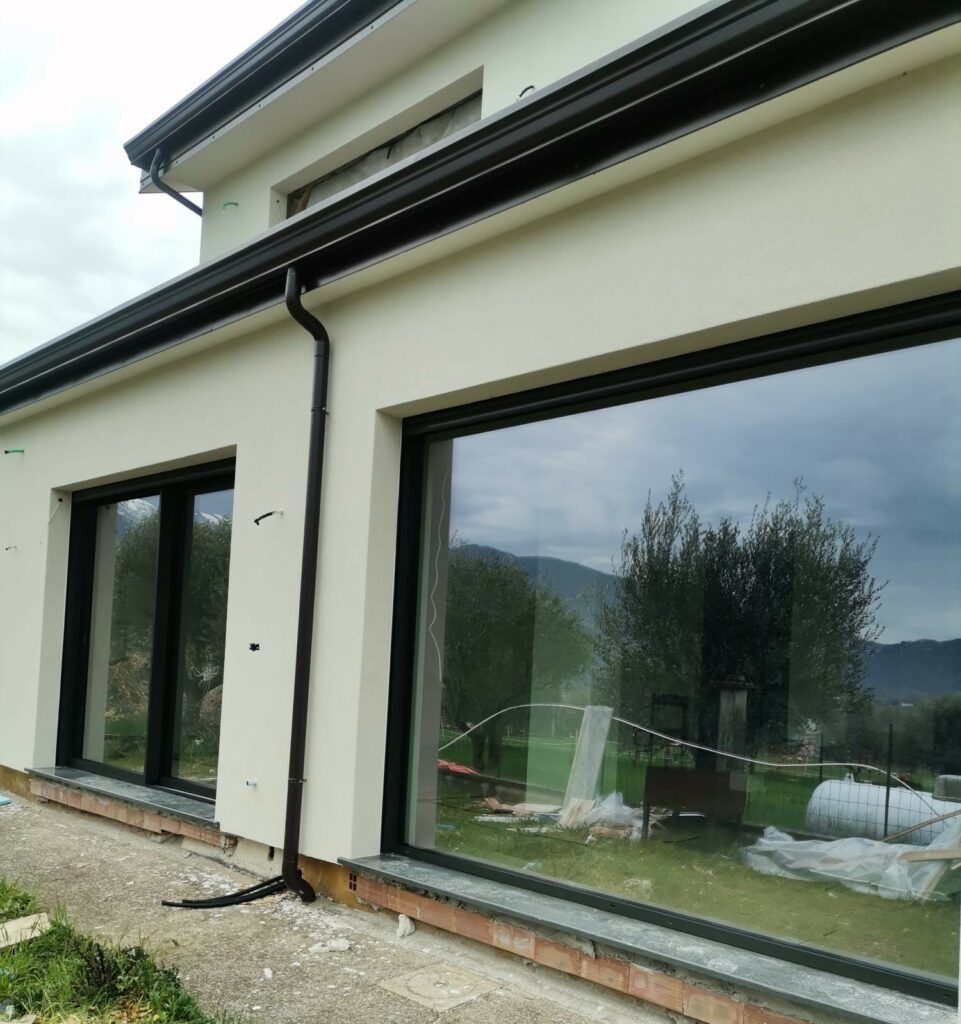 Esterno di una casa moderna con ampie finestre e cornici nere, che riflettono un cortile e un cielo nuvoloso. Pareti in stucco bianco, grondaie nere.