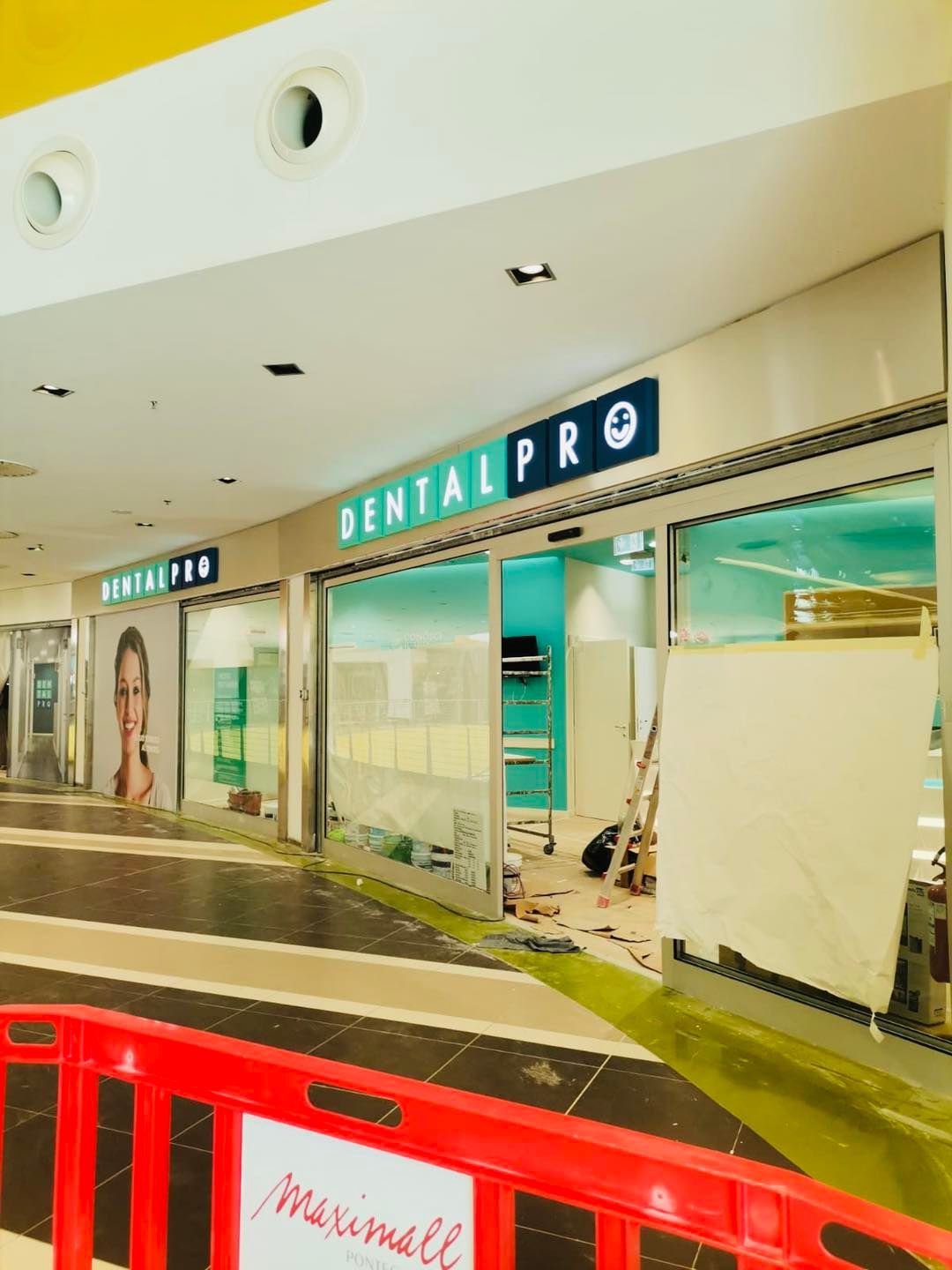Vetrina di Dental Pro in un centro commerciale, in costruzione. Cartello e interni azzurri visibili. Barriera rossa all'ingresso.