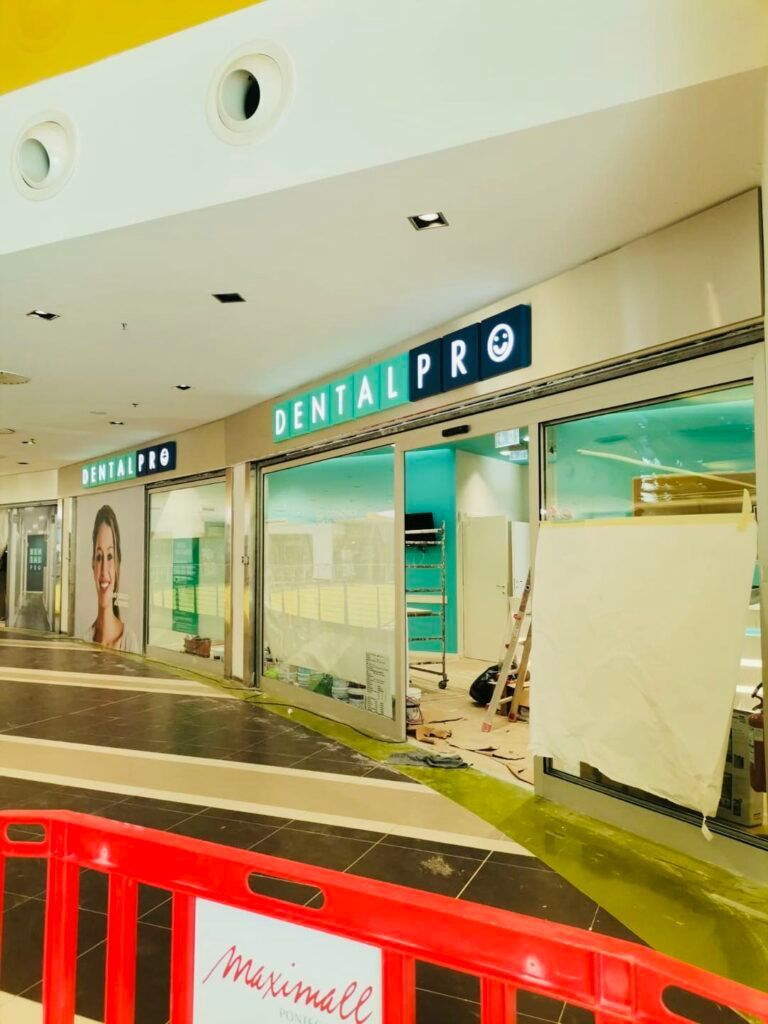 Clinica Dental Pro in un centro commerciale, con una barriera rossa all'ingresso. La segnaletica è illuminata in verde.