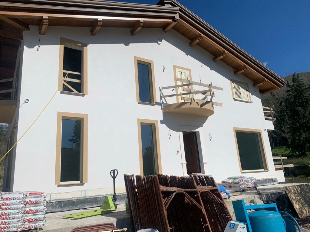 Casa a due piani in costruzione con pareti in stucco bianco, infissi marroni e un piccolo balcone. I materiali da costruzione sono già presenti sul davanti.