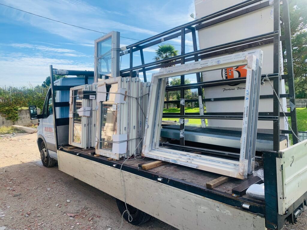 Un camion con pianale carico di infissi bianchi, parcheggiato all'aperto in una giornata di sole. Alcuni finestrini sono impilati, altri appoggiati al bordo.