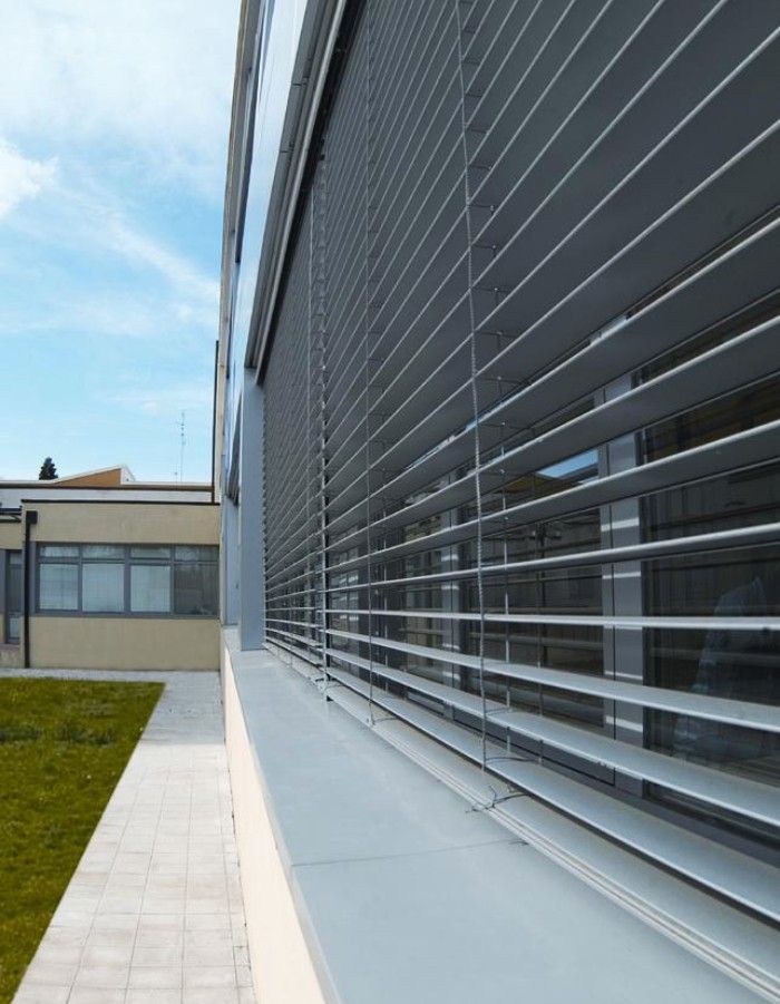 Edificio moderno con tende da sole orizzontali a stecche sopra grandi finestre; comprende un piccolo edificio con finestre laterali.
