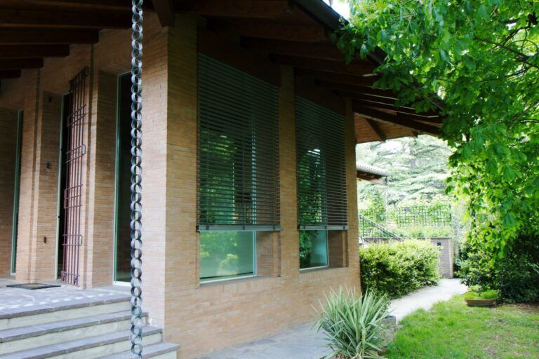 Edificio marrone chiaro con due finestre coperte da persiane. Una catena per la pioggia è appesa sul lato sinistro. Il fogliame verde circonda l'edificio.