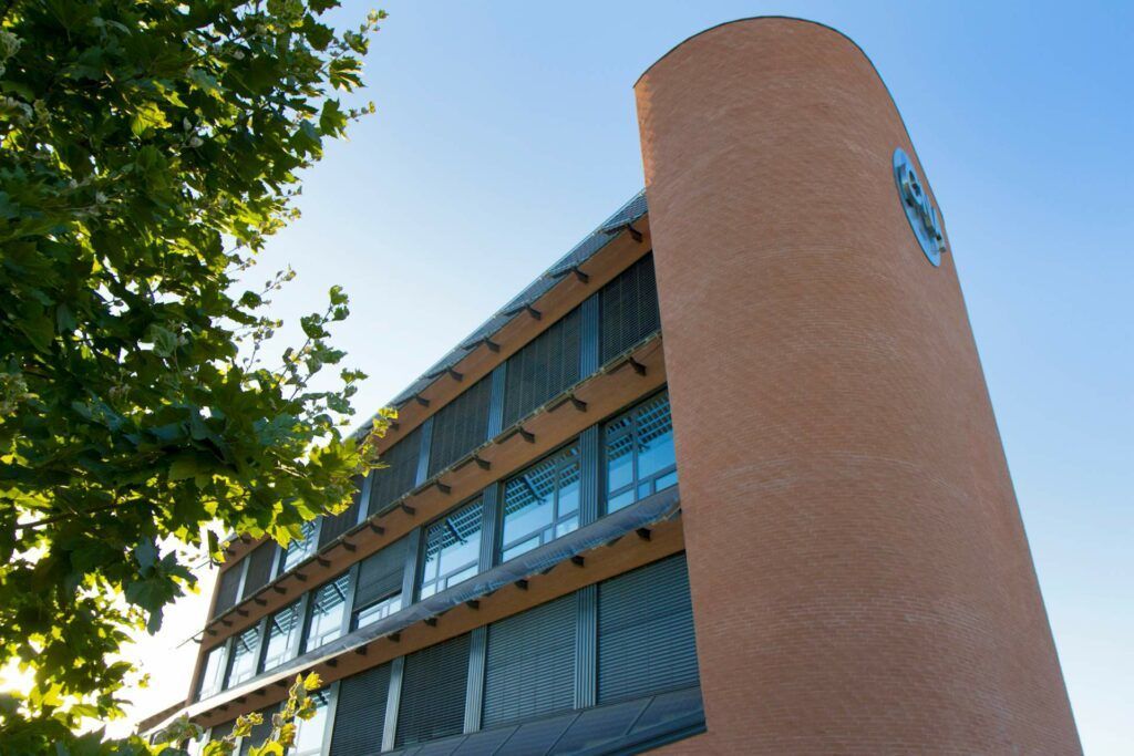 Edificio in mattoni con una torre curva, cielo azzurro sullo sfondo. La luce del sole filtra attraverso i rami degli alberi in primo piano.