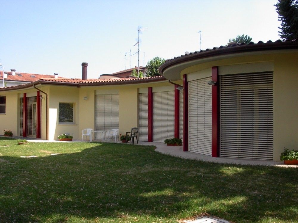 Edificio basso, di colore giallo chiaro con accenti rossi, caratterizzato da sezioni curve, finestre con persiane chiuse e un prato verde.