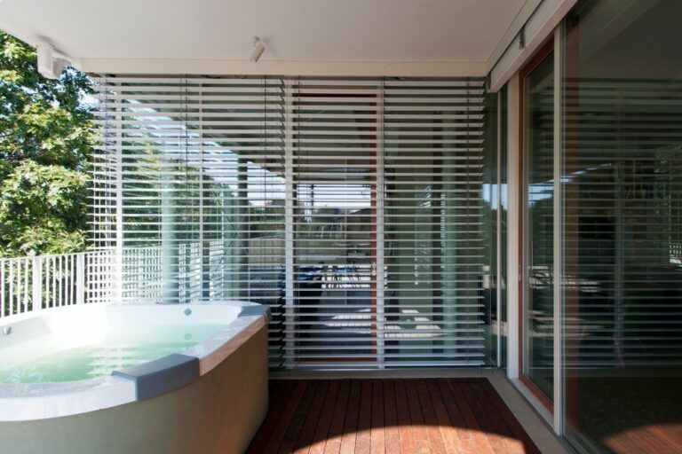 Una vasca idromassaggio su una terrazza in legno con una tenda a doghe e una porta scorrevole in vetro; all'esterno si vede il fogliame verde.