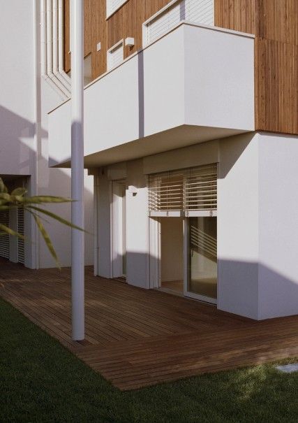 Vista esterna di un edificio moderno con pareti bianche, dettagli in legno e una terrazza in legno. La luce del sole proietta ombre sulla facciata.