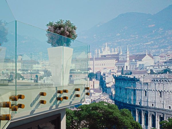 Un balcone in vetro con una fioriera bianca che si affaccia sul paesaggio urbano e sulle montagne sotto un cielo azzurro velato.