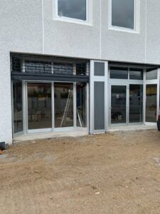 Vista esterna di un edificio con ampie porte e finestre in vetro. L'edificio ha una facciata di colore chiaro e sorge su un terreno sterrato.