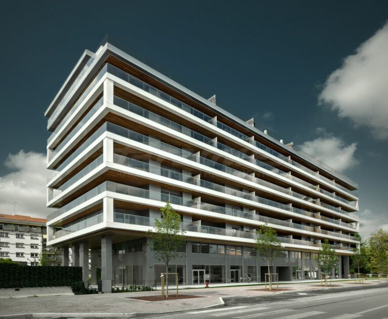 Edificio moderno a più piani con linee orizzontali, balconi in vetro e pilastri in cemento. La struttura si affaccia su una strada, con alberi e un cielo parzialmente nuvoloso sullo sfondo.