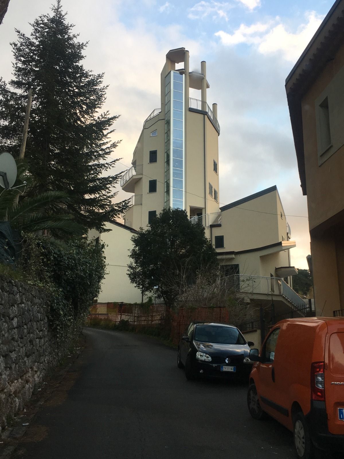 In fondo a una stretta strada si erge un alto edificio color crema con un vano ascensore in vetro, davanti al quale sono parcheggiate delle auto.