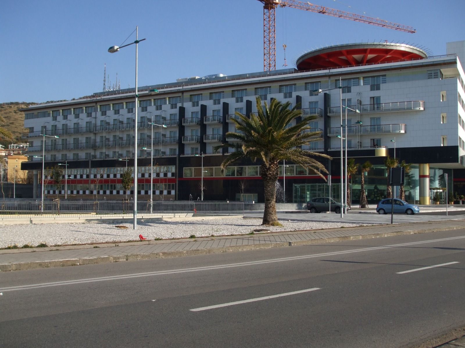 Un moderno edificio a più piani con una struttura circolare con la sommità rossa sul tetto, una gru da costruzione e palme in un ambiente soleggiato.