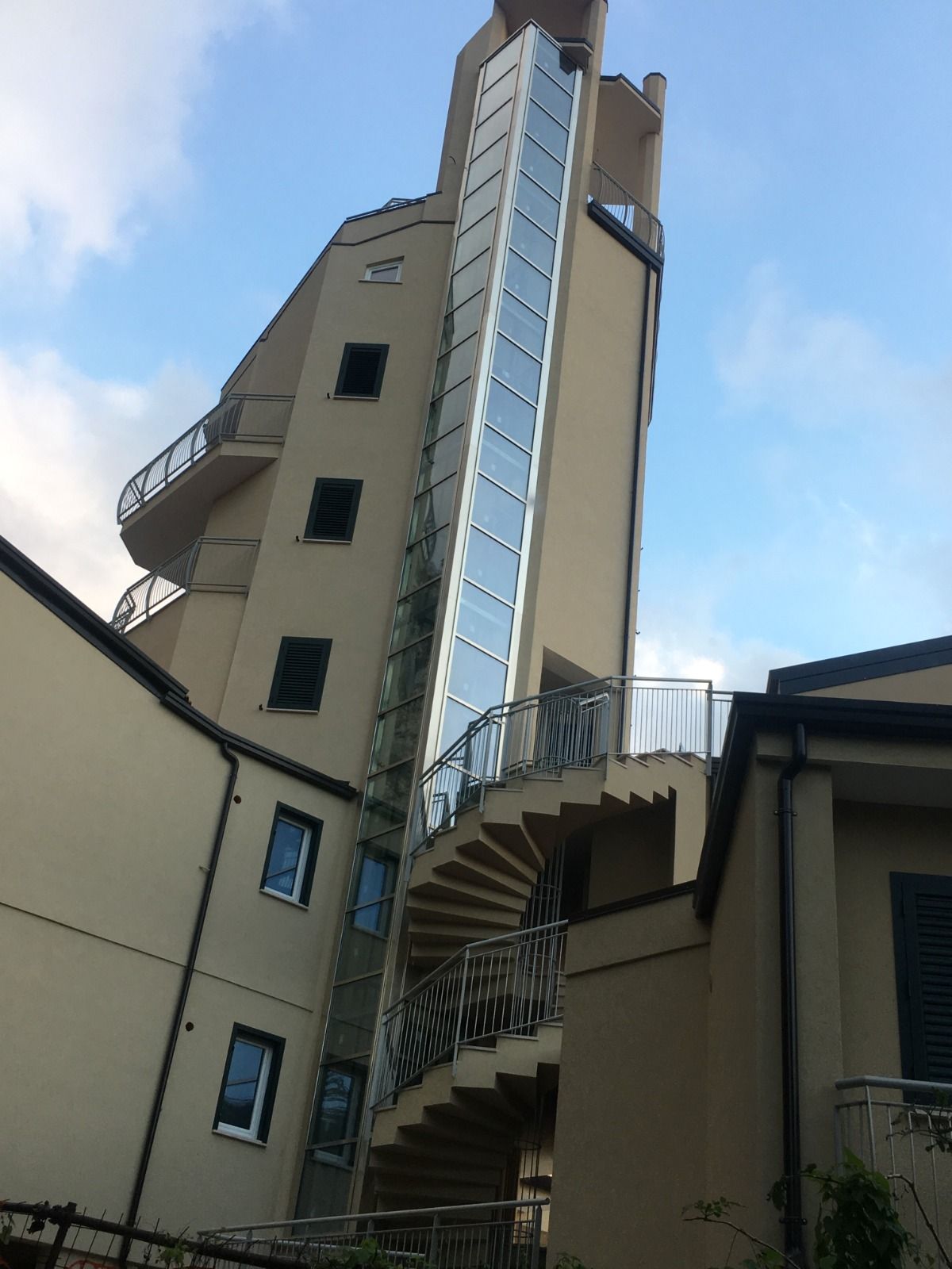 Un alto edificio beige con un ascensore in vetro e una scala a chiocciola esterna, che si staglia contro un cielo parzialmente nuvoloso.