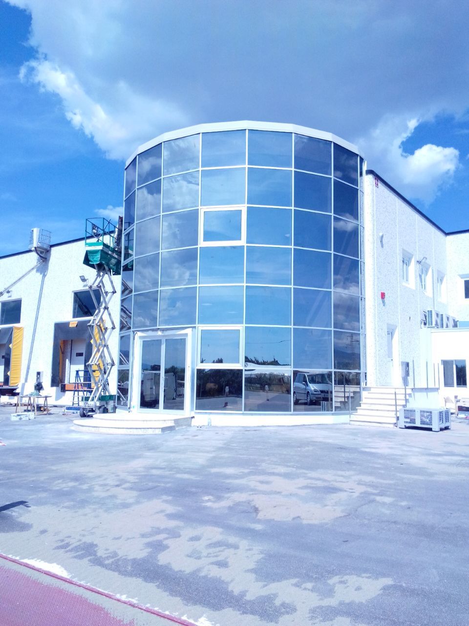 Edificio moderno con una facciata curva in vetro che riflette il cielo azzurro. Vicino all'edificio è presente una piattaforma elevatrice.