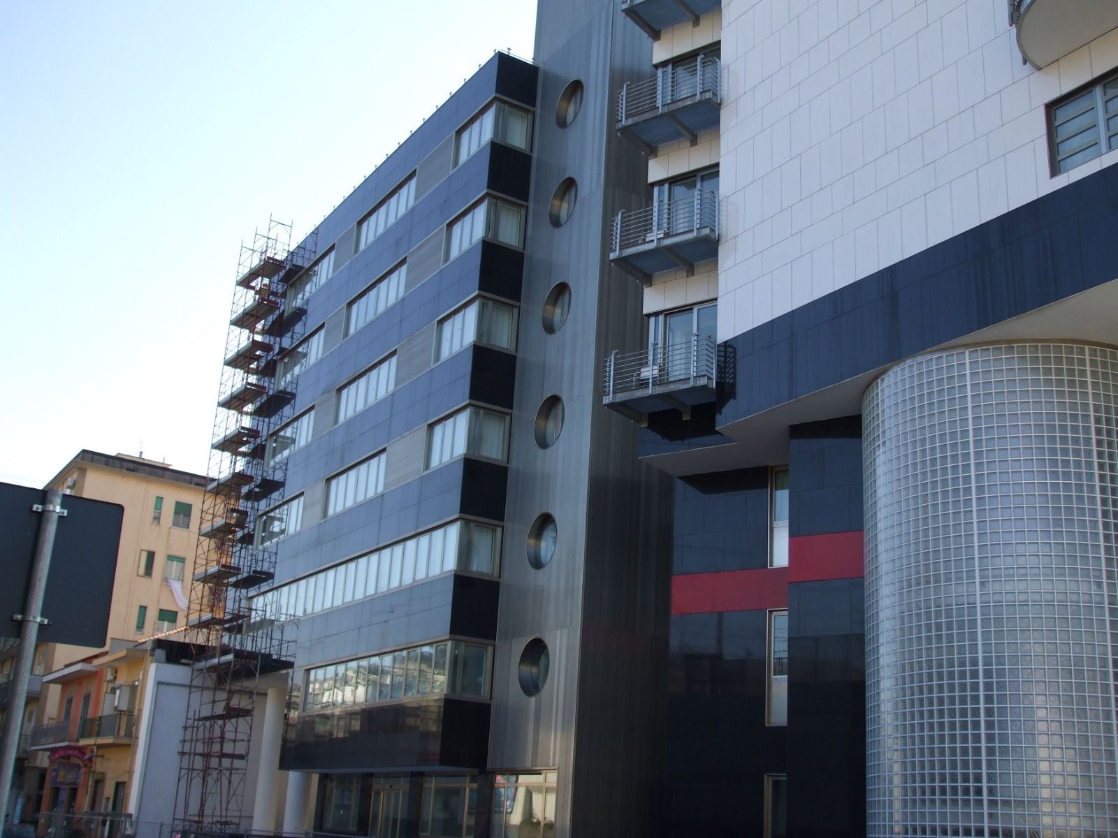 Esterno di un edificio moderno con pannelli scuri riflettenti e finestre rotonde. Impalcature visibili lateralmente.