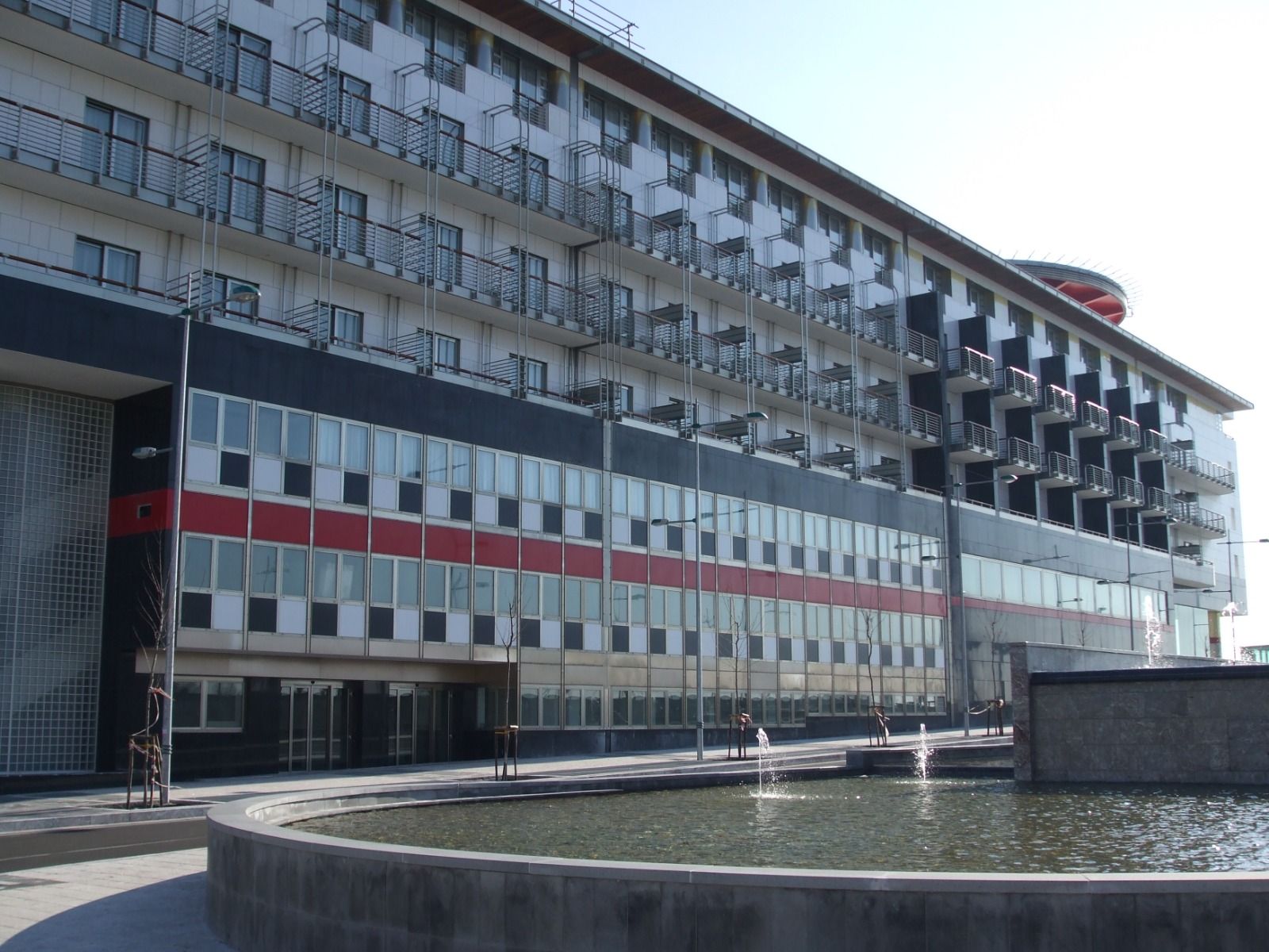 Un lungo edificio a più piani con balconi, fasce alternate nere, rosse e bianche. Una fontana si trova in primo piano.
