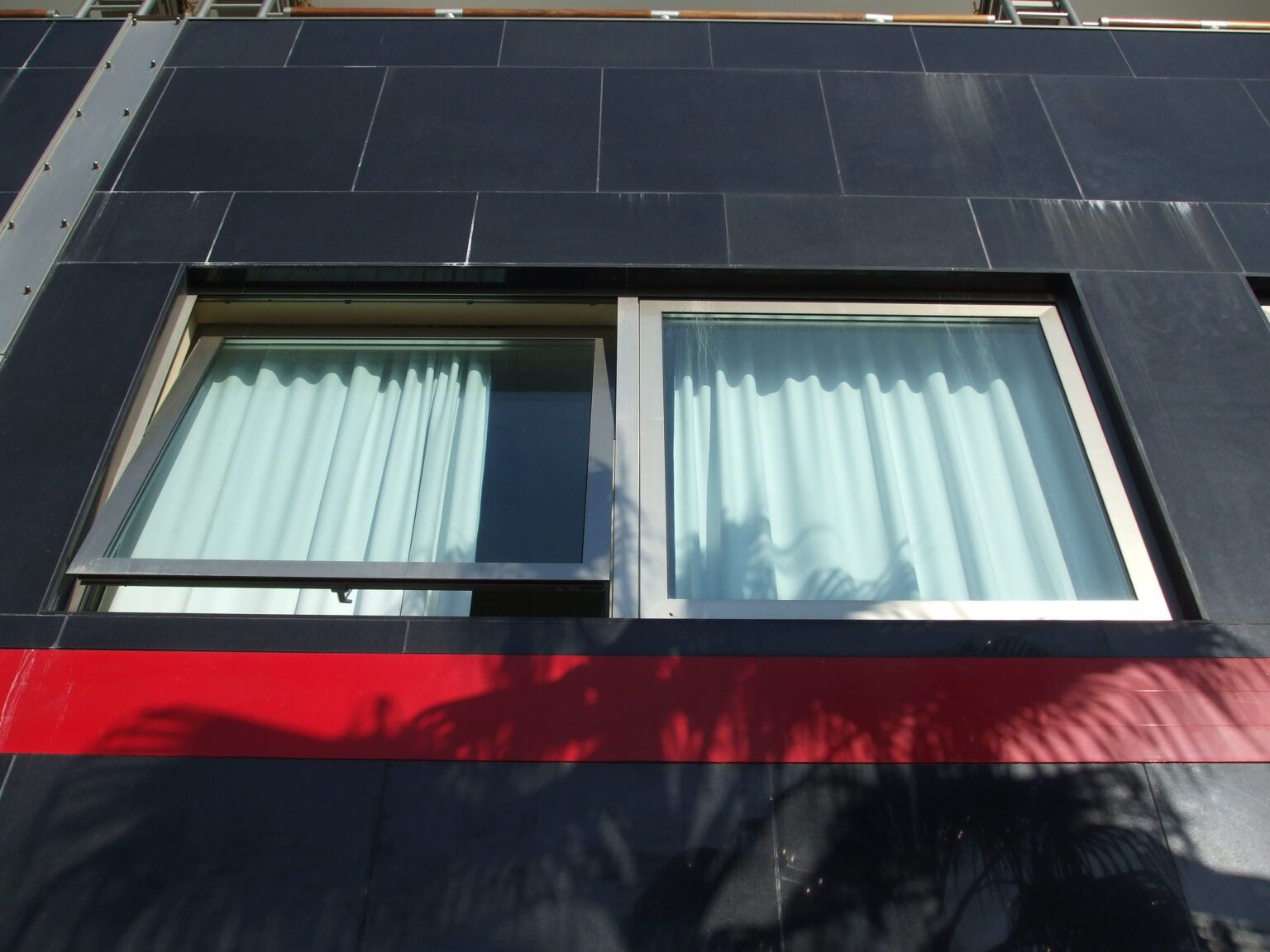 Due finestre con cornici bianche e tende azzurre su un edificio scuro con una striscia rossa.