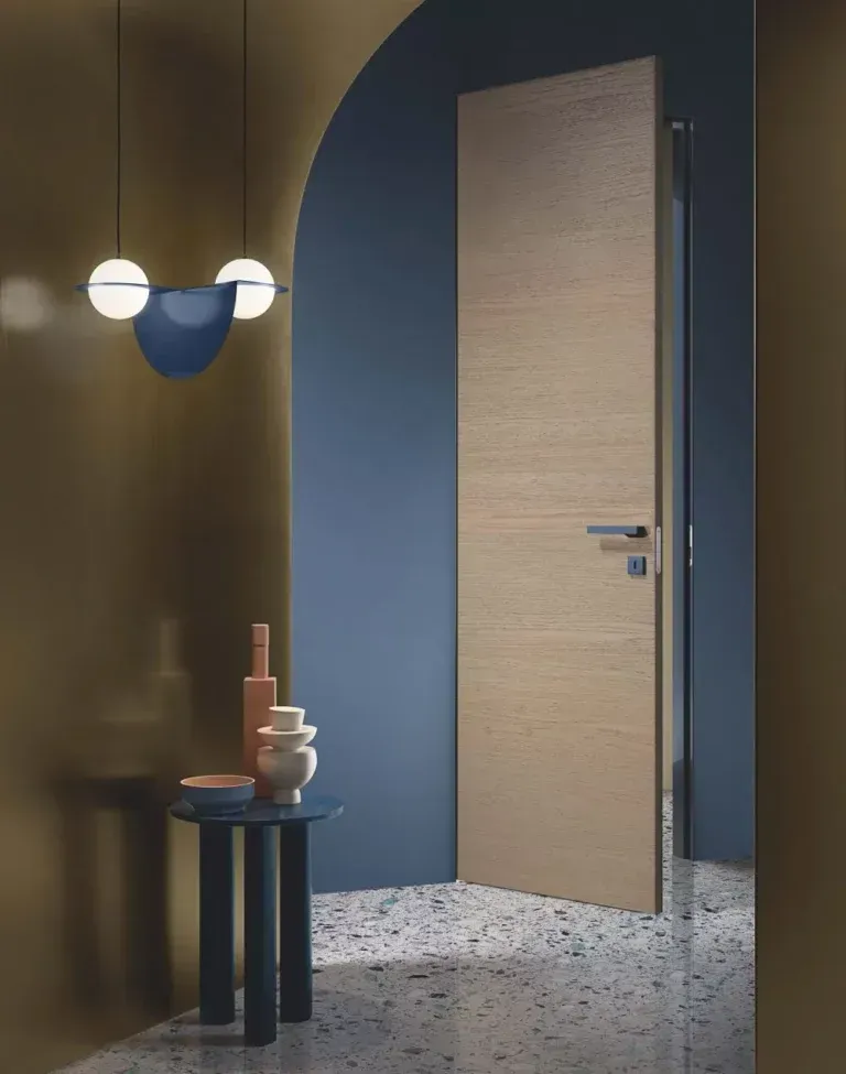 Un interno moderno caratterizzato da una porta in legno chiaro contro una parete blu, con un tavolino decorativo e un'elegante lampada a sospensione.