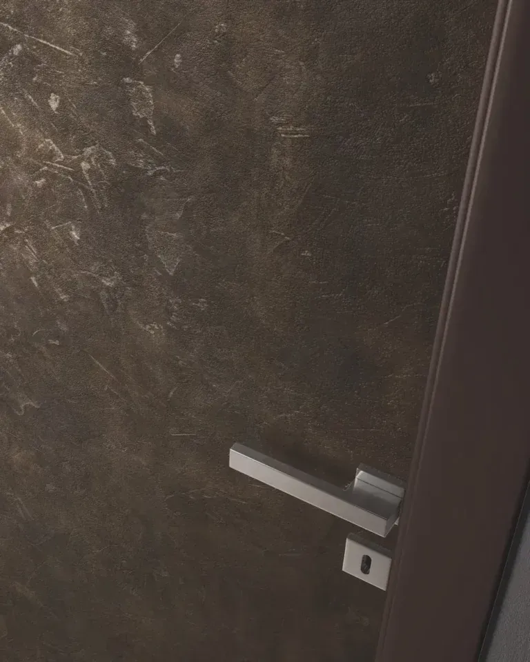 Primo piano di una porta scura e ruvida con maniglia e serratura argentate. Sulla destra è visibile uno stipite marrone.