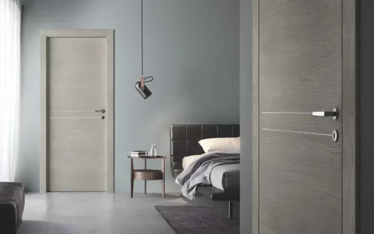 Interno di una camera da letto con due porte in legno chiaro, un letto e un comodino contro una parete grigio-blu.
