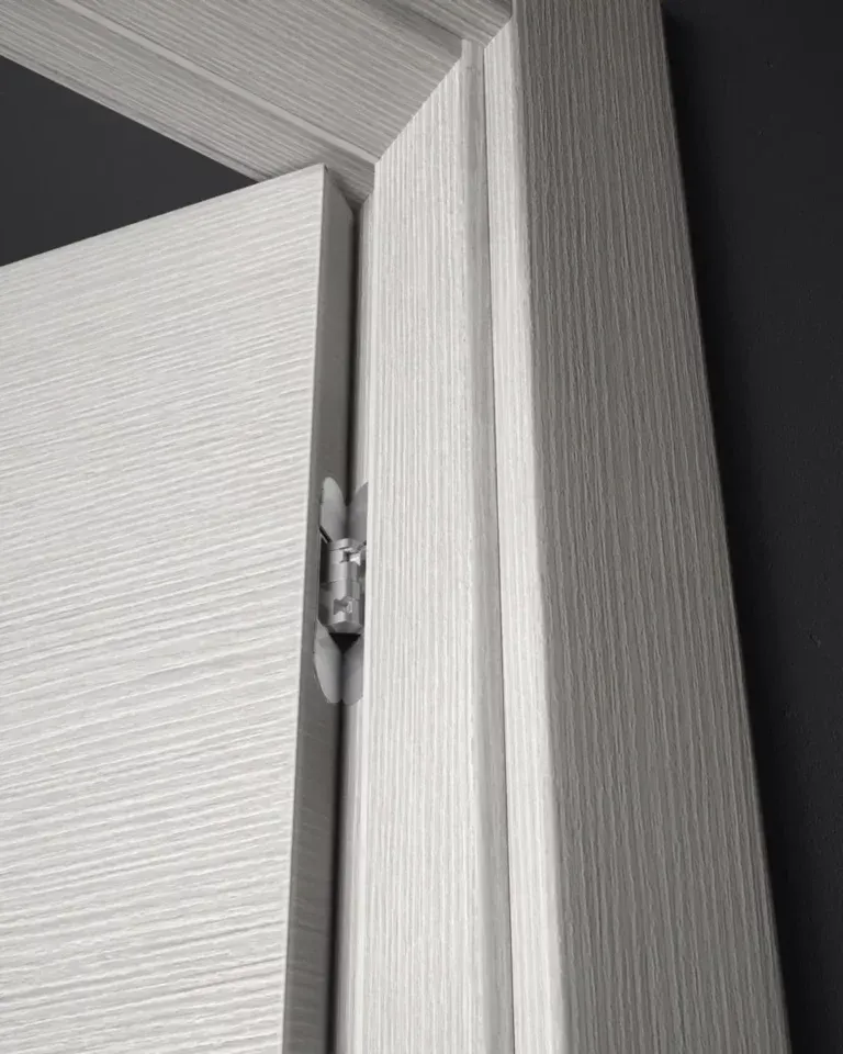 Primo piano di una porta e di un telaio in legno bianco, che rivelano una cerniera argentata. La porta e il telaio presentano una venatura lineare e strutturata.