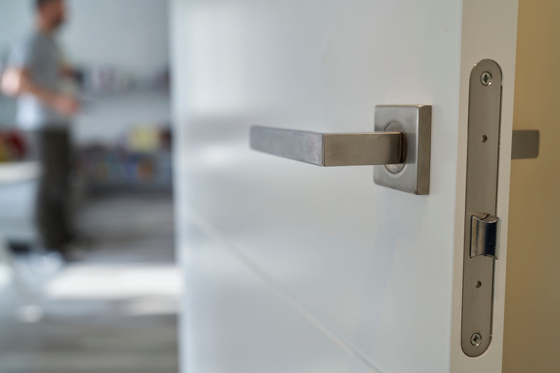 Primo piano di una porta bianca con maniglia e serratura argentate; sullo sfondo è sfocata una persona.