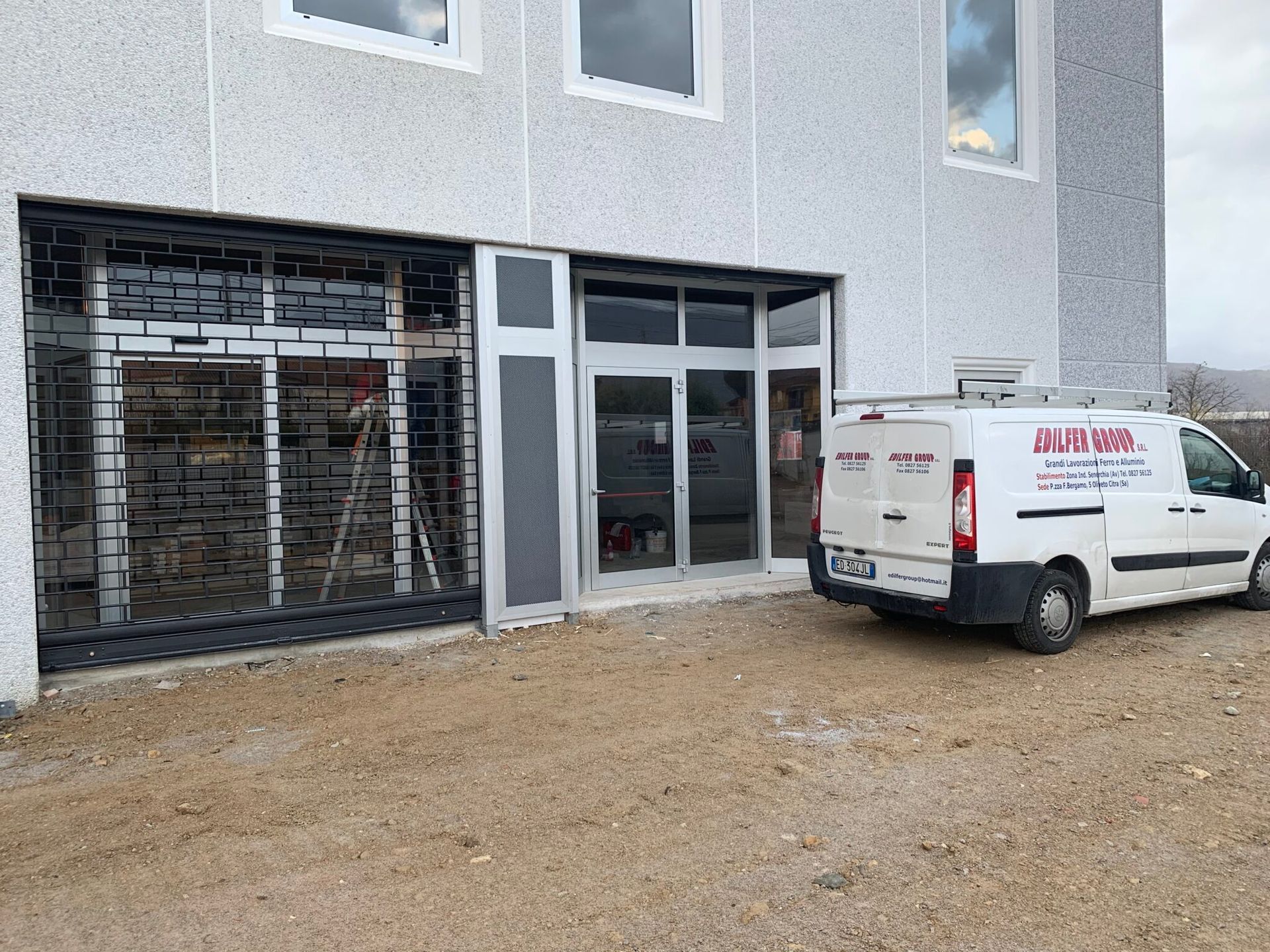 Esterno di un edificio con cancello di sicurezza chiuso e ingresso con porta a vetri. Un furgone bianco con il logo aziendale è parcheggiato accanto all'ingresso.
