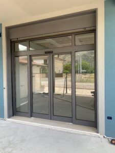 Porte scorrevoli automatiche in vetro grigio con telai grigi, inserite in un edificio esterno azzurro.