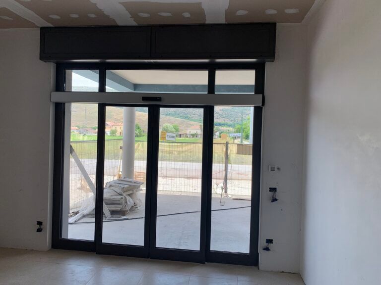 Porte scorrevoli in vetro con telai scuri, meccanismo di apertura automatizzato e vista su un'area esterna con recinzione e paesaggio verde.