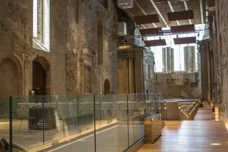Vista interna di uno storico edificio in pietra con moderne ringhiere in vetro e pavimenti in legno, che ospita una mostra.