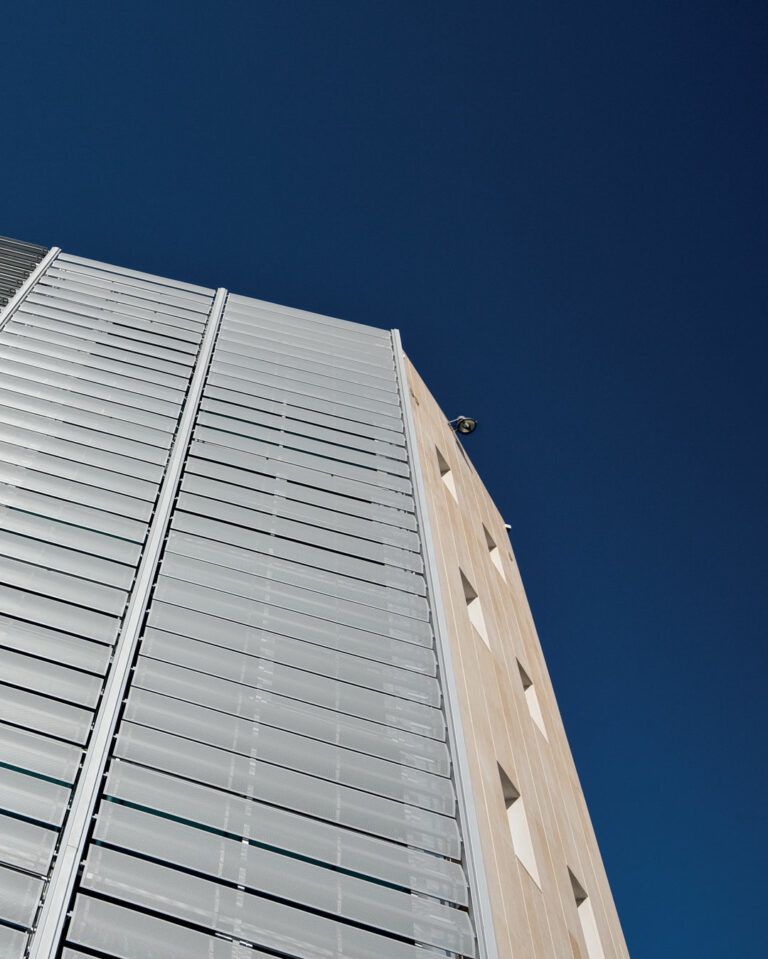 Vista dal basso di un alto edificio moderno contro un cielo azzurro terso. Un lato presenta pannelli chiari e supporti metallici; l'altro, una facciata testurizzata color marrone chiaro con aperture per finestre.