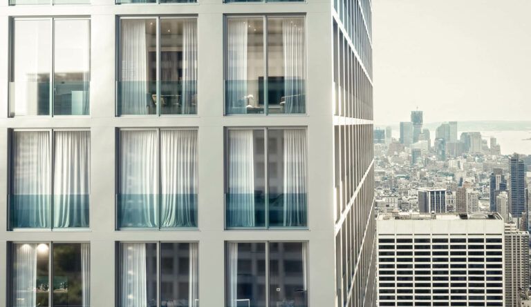 Primo piano dell'esterno di un moderno grattacielo con ampie finestre; sullo sfondo è visibile lo skyline della città.