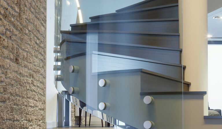 Scala moderna con gradini grigi, pannello in vetro e parete in mattoni sulla sinistra. Fissaggi metallici fissano il vetro alla scala.