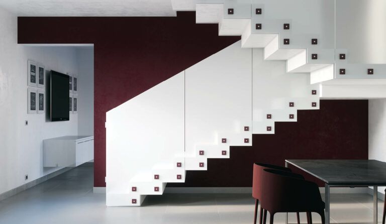 Una moderna scala bianca sospesa con parete bordeaux, tavolo da pranzo e uno scorcio di soggiorno.