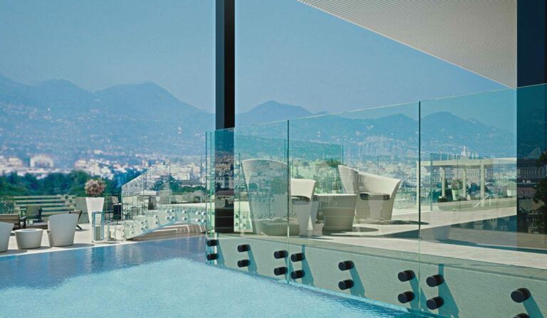 La piscina a sfioro con parapetto in vetro si affaccia sulla città e sulle montagne. Sul patio sono presenti delle sedie a sdraio bianche.