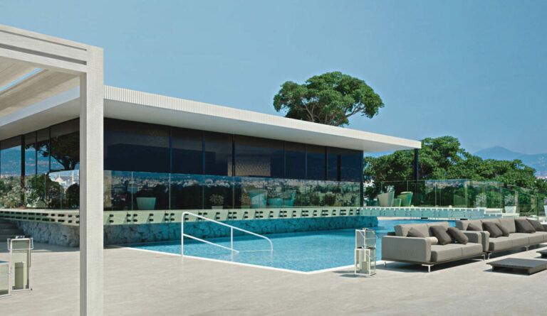 Edificio moderno con piscina e posti a sedere all'aperto; ampie finestre riflettono il cielo azzurro.