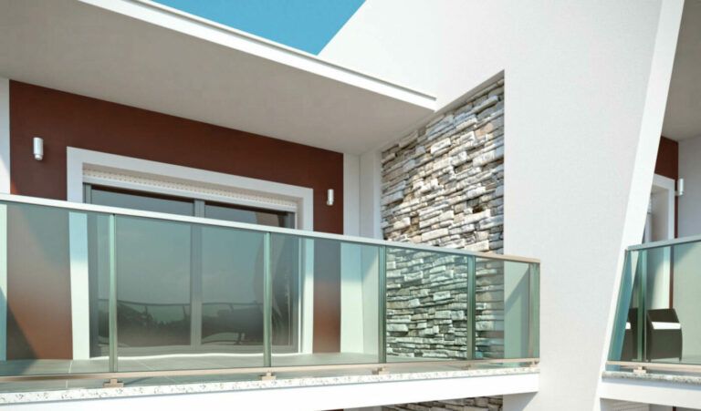 Balcone con ringhiera in vetro, parete decorativa marrone e muro in pietra. Esterno dell'edificio bianco con cielo azzurro visibile.