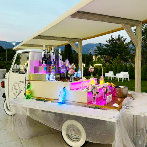 Ape Piaggio bianca trasformata in bar mobile. Bevande, fiori e bottiglie esposti sotto una tettoia durante un evento all'aperto.